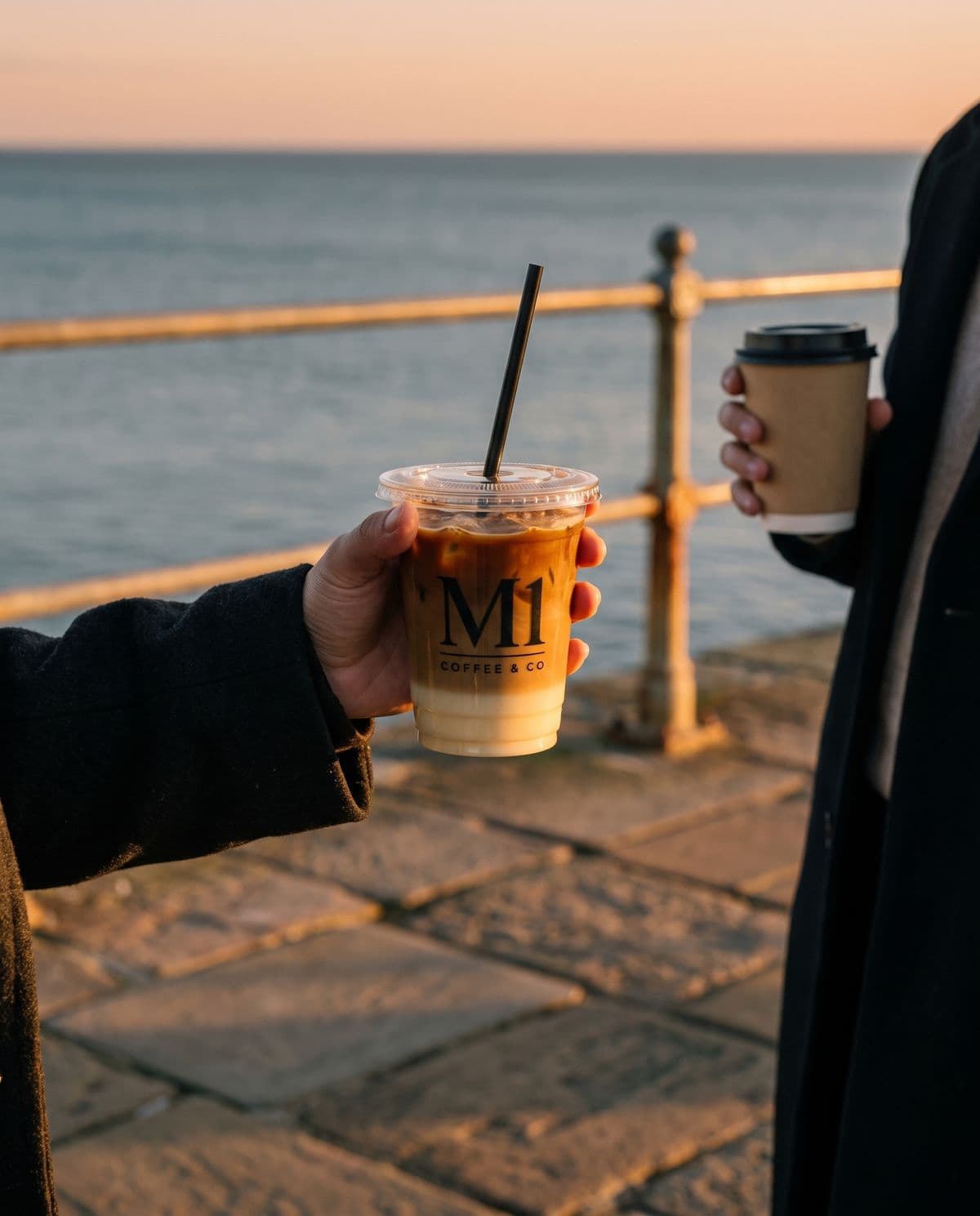 A hand holding an iced M1 coffee on the New Brighton promenade at golden hour